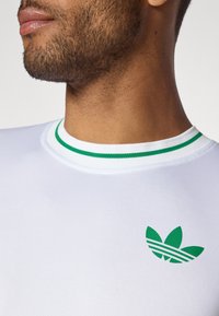 Fehér sportpóló kerek nyakkal, zöld csíkokkal. Zöld logó a mellrészen három levélformával és finom vonalakkal.