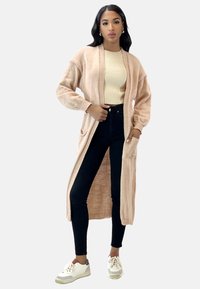 Cardigan lungo con apertura anteriore in morbido maglione rosa chiaro con dettagli a costine. Indossato sopra un top beige, abbinato a jeans neri a vita alta e sneakers bianche.