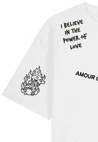 T-shirt blanc avec texte noir "JE CROIS EN LE POUVOIR DE L’AMOUR" et art linéaire de dés en flammes sur la manche.