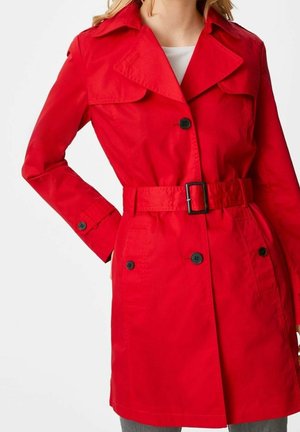 Kvinde iført en knaldrød trenchcoat med bælte og sorte knapper, stående mod en ensfarvet hvid baggrund.