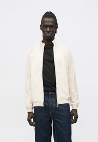 Lys beige bomberjakke med glidelås, strukturert diamantmønster, ribbede mansjetter og krage; brukt over en svart skjorte og mørke denim bukser.