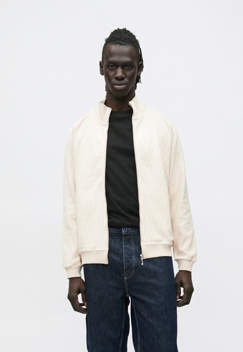 Lys beige bomberjakke med glidelås, strukturert diamantmønster, ribbede mansjetter og krage; brukt over en svart skjorte og mørke denim bukser.