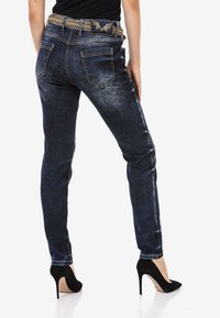 Jean en denim bleu foncé avec une finition brillante, design ajusté et ceinture accentuée de doré. Détails de couture et deux poches arrière visibles.