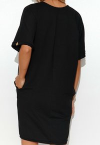 Schwarzes kurzärmeliges Kleid mit lockerer Passform, rundem Ausschnitt, seitlichen Taschen und einem sichtbaren goldenen Knopfakzent am Ärmel.