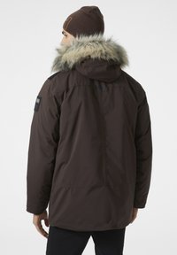 Hnědá parka bunda s kapucí lemovanou kožešinou, s hladkým povrchem, černými akcenty na ramenech a logem na rukávu.