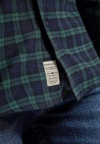 Zielona i granatowa koszula w kratę z teksturowanej tkaniny. Posiada metkę z napisem "CROSS JEANS" w białym kolorze, wyszytą wzdłuż dolnej krawędzi.