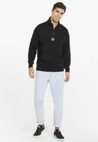 Felpa nera con zip e collo alto, con tasca frontale e un piccolo logo bianco. Abbinata a jeans azzurri chiari e sneakers nere.