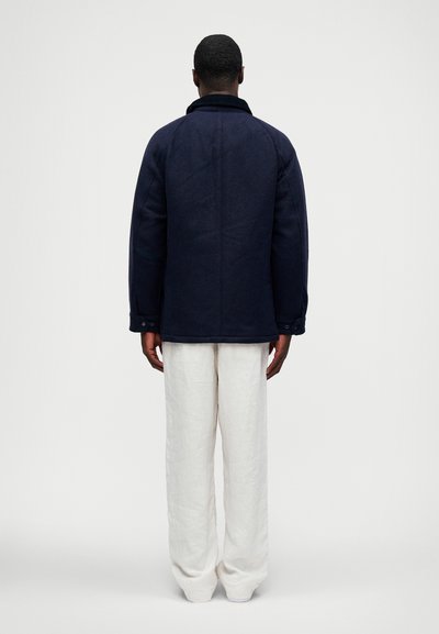 Homem de pé, de costas, vestindo um casaco azul-marinho escuro e calças brancas largas, contra um fundo claro e simples.