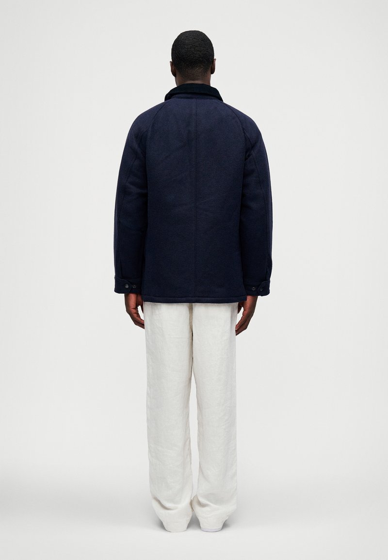 Homme se tenant de dos, portant une veste bleu marine foncé et un pantalon blanc ample, sur un fond clair et uni.