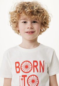 Biała bawełniana koszulka z czerwoną grafiką, na której znajduje się napis "BORN TO" oraz okrągłe wzory kół. Model ma kręcone blond włosy.
