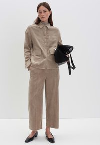 Femme en veste en velours côtelé beige et pantalons larges, tenant un sac en filet noir, portant des chaussures à petit talon noires sur un fond blanc.