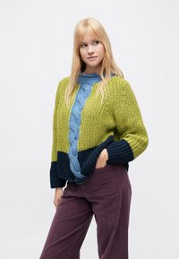 Storas megztinis su pynimu, kuriame yra žalių, mėlynų ir tamsiai mėlynų skyrių. Laisvas siluetas su ribinėmis rankogaliais ir apačia.