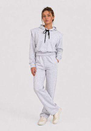 Donna in piedi su sfondo semplice indossa una felpa con cappuccio grigio chiaro con coulisse nere, pantaloni della tuta abbinati e sneakers beige.
