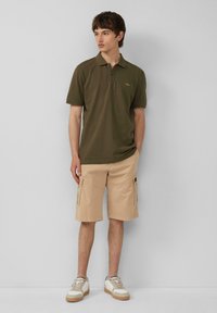 Olivgrünes Polohemd mit Kragen und kleinem Logo, kombiniert mit beigen Cargo-Shorts mit Seitentaschen und weißen Sneakers mit braunen Akzenten.