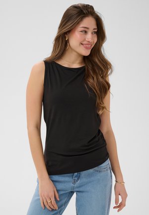 Jeune femme aux longs cheveux bruns portant un haut noir sans manches et un jean bleu clair, souriante et regardant sur le côté.