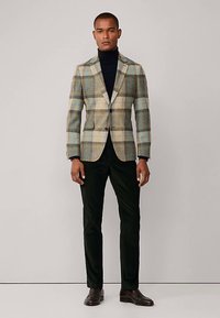 Marynarka w kratę w kolorach beżowym, niebieskim i brązowym z dwoma guzikami, zestawiona z czarnym golfem i ciemnozielonymi spodniami. Brązowe buty dopełniają stylizację.