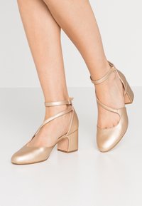 Metalliska guldblockklackade skor med rund tå, ankelremmar och urtagna sidor. Slät textur och glänsande ytfinish.
