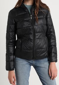 Svart puffjacka med kviltat mönster, dragkedja framtill, ståkrage och elastisk midja. Tillverkad av glänsande, lätt material.