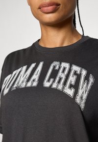 Camiseta de algodón negra con cuello redondo, que presenta un texto desgastado en blanco en negrita que dice "PUMA CREW" en el pecho. Textura suave.