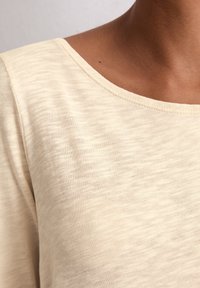 Haut en tricot texturé beige clair, avec un col large et arrondi et des manches courtes, présentant une finition douce et légèrement chinée.