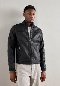 Jachetă din piele neagră cu detalii ondulate pe umeri și talie, fermoar frontal și buzunare, design ajustat, asociată cu pantaloni gri deschis.