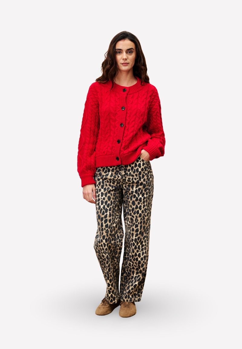 Cardigan rouge en maille torsadée avec des boutons noirs, associé à un pantalon ample en imprimé léopard et des chaussures marron. Design simple et tissu texturé.