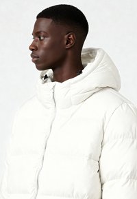 Giacca imbottita bianca con collo alto e coulisse, con chiusura a zip e texture trapuntata, indossata da una persona vista di lato.