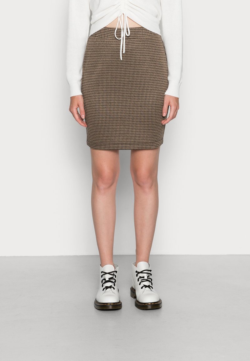 Jupe crayon en pied-de-poule marron et crème, en tissu texturé, longueur au-dessus du genou. Associée à des bottes blanches à lacets noirs.