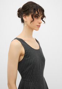 Hame vestido sin mangas en tela gris oscuro con rayas blancas verticales. Cuerpo ajustado que se abre en una falda acampanada. Diseño simple y elegante. Aretes de aro dorados.