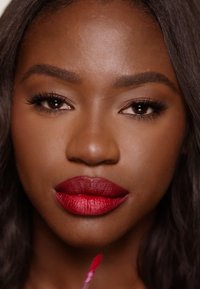 Roter flüssiger Lippenstift, auf voller, strukturierter Lippen aufgetragen. Der satte Farbton kontrastiert mit glatter, dunkler Haut. Lange Wimpern umrahmen breite, mandelförmige Augen.