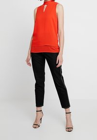 Červená bezrukávová blůza s výstřihem ve tvaru klíčové dírky, spárovaná s černými upnutými kalhotami s bočním švem. Černé sandály na vysokém podpatku dokončují vzhled.