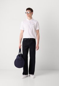 Hvit polo skjorte, mørke jeans og hvite sneakers. Holder en rund marineblå veske med håndtak. Tilbehør inkluderer firkantede solbriller.