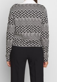 Long-sleeve black and white blouse featuring a zigzag pattern, with a fitted silhouette and round neckline. Paired with black trousers.