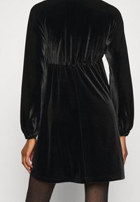 Černé sametové šaty s dlouhými, nabíranými rukávy. Nařasený pas, silueta A-linie a hladká textura. Výrazný lesk na látce.