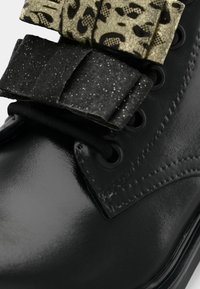 Scarpa in pelle nera lucida con fiocchi in tessuto nero e oro a motivo strutturato attaccati vicino ai lacci.