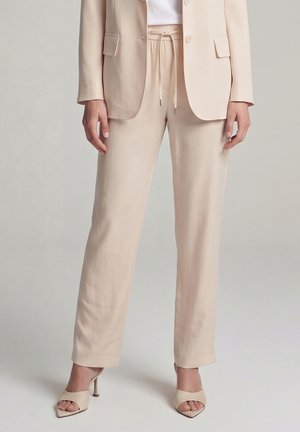 Woman wearing beige tailored trousers with drawstring waist, matching blazer, and open-toe heeled sandals, standing against neutral background.