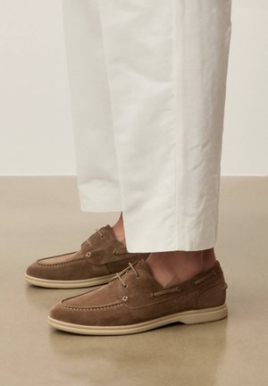 Mocassins en daim marron à lacets portés avec un pantalon blanc à jambes droites sur un sol beige lisse.