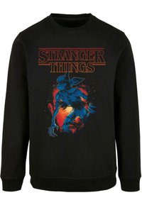 Crna dukserica s vibrantnom grafikom plavog stvorenja među svijetlim crvenim i narančastim naglascima, uz natpis "Stranger Things" iznad.