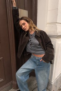 Brown suede jacket, gray ribbed top with buttons, and light blue baggy jeans. Background features a brown door and stone wall.