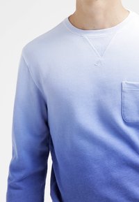Hombre vistiendo una sudadera de manga larga tipo crewneck con un degradado que va de lavanda claro a azul profundo y un bolsillo en el lado izquierdo del pecho.