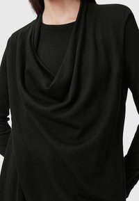 Femme portant un haut noir à manches longues avec un col bénitier drapé, vue du cou jusqu'aux hanches sur un fond clair uni.