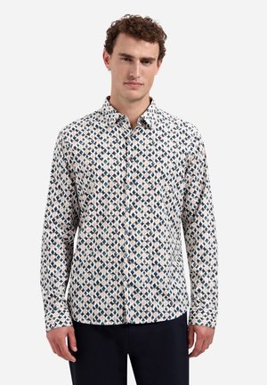 Mann trägt langärmliges Hemd mit Knöpfen und geometrischem Muster in Blau, Beige und Weiß, kombiniert mit dunkler Hose, steht vor weißem Hintergrund.