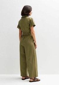 Mono verde oliva con mangas cortas, cinturilla ceñida, piernas anchas y puños enrollados. Se lleva con sandalias marrones, mostrado desde la parte de atrás.