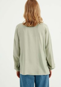 Blusa verde chiaro a maniche lunghe con vestibilità rilassata, caratterizzata da un collo rotondo e polsini leggermente arricciati, abbinata a jeans in denim blu.