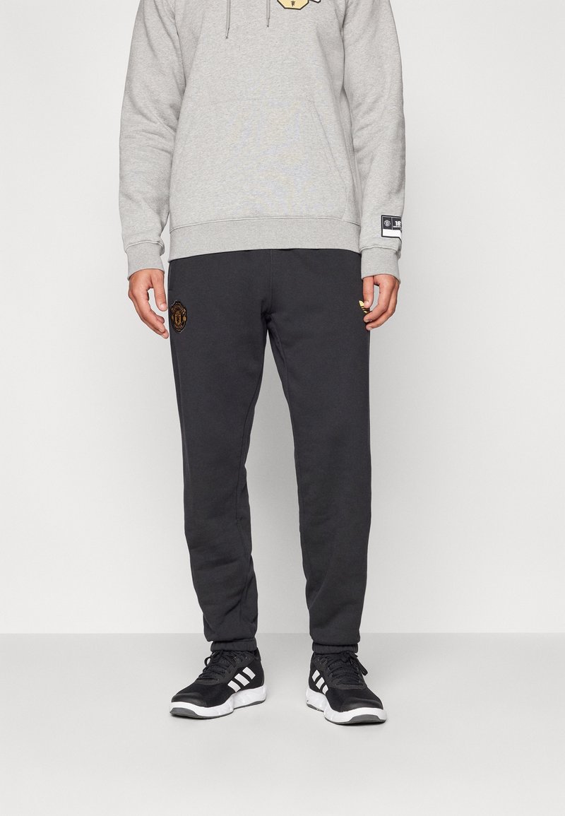 Tummanharmaat collegehousut, joissa vasemmassa reidessä kirjailtu logo, yhdistetty vaaleanharmaaseen huppariin, jossa on graafinen kuosi ja kiristysnauha. Mustat lenkkarit.