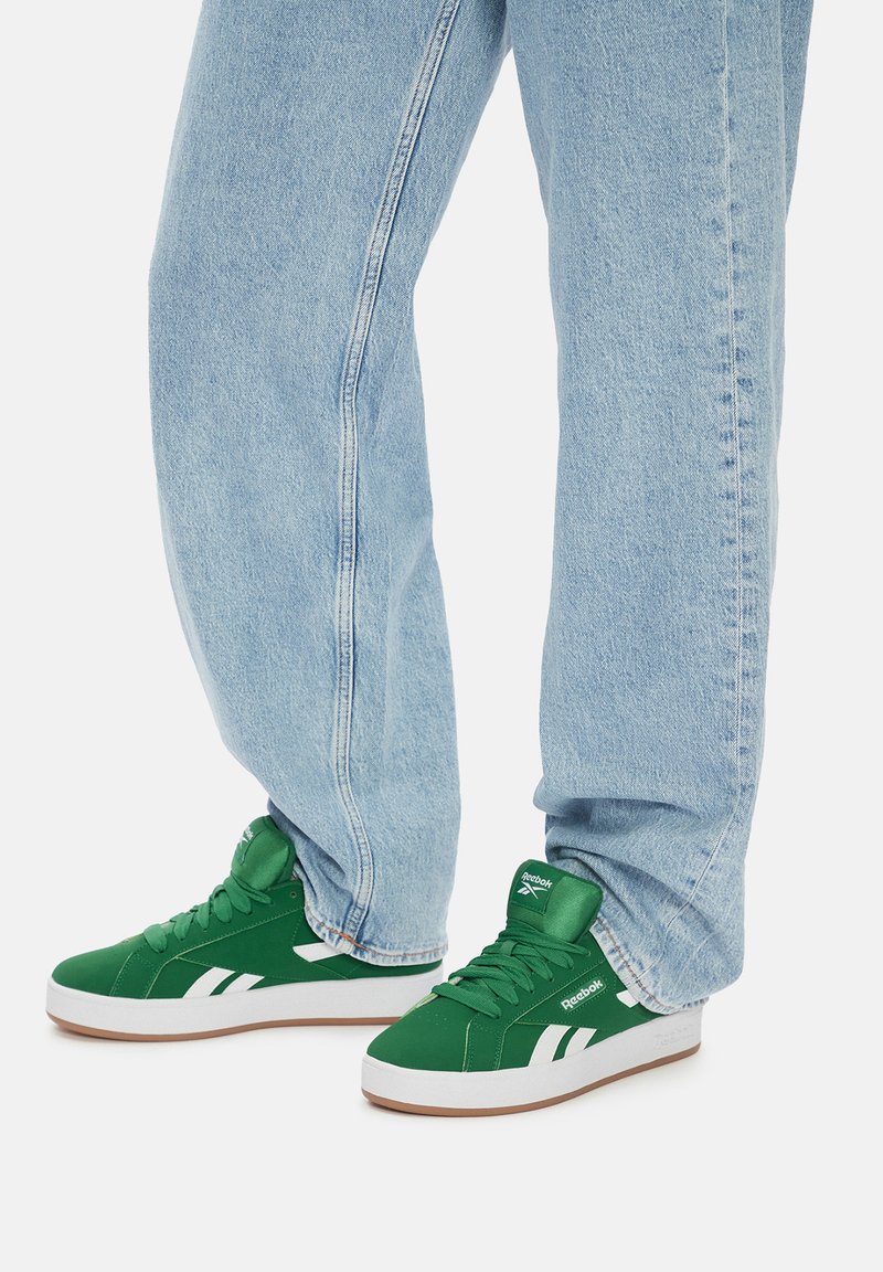Zielone trampki z białymi akcentami, z wyściełanym kołnierzem, okrągłym noskiem i teksturowaną gumową podeszwą, zestawione z jasnoniebieskimi dżinsami.