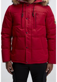 Veste matelassée rouge avec capuche bordée de fourrure, fermeture éclair, deux poches avant et motif matelassé horizontal. Détails noirs et logo.