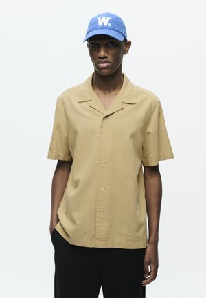 Korte mouwen, lichtbruine knoopshirt met een kraag en subtiele textuur, gecombineerd met een blauwe pet met een wit logo.