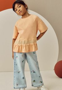 Helles oranges Kurzarm-Oberteil mit einem Spitzenblumenbesatz; kombiniert mit hellblauen, weit geschnittenen Jeans mit bunten Blumenstickereien.