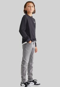 Svart långärmad skjorta med vit logotyp, ihop med ljusgrå tajta jeans och svart-vita sneakers, visad mot en neutral bakgrund.